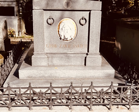 Zdjęcie grobu rodziny Zawiejskich. Na płycie biała płaskorzeźba twarzy Jezusa, złoty napis z nazwiskiem zmarłych. Wokół kute, niskie, czarne, metalowe ogrodzenie.