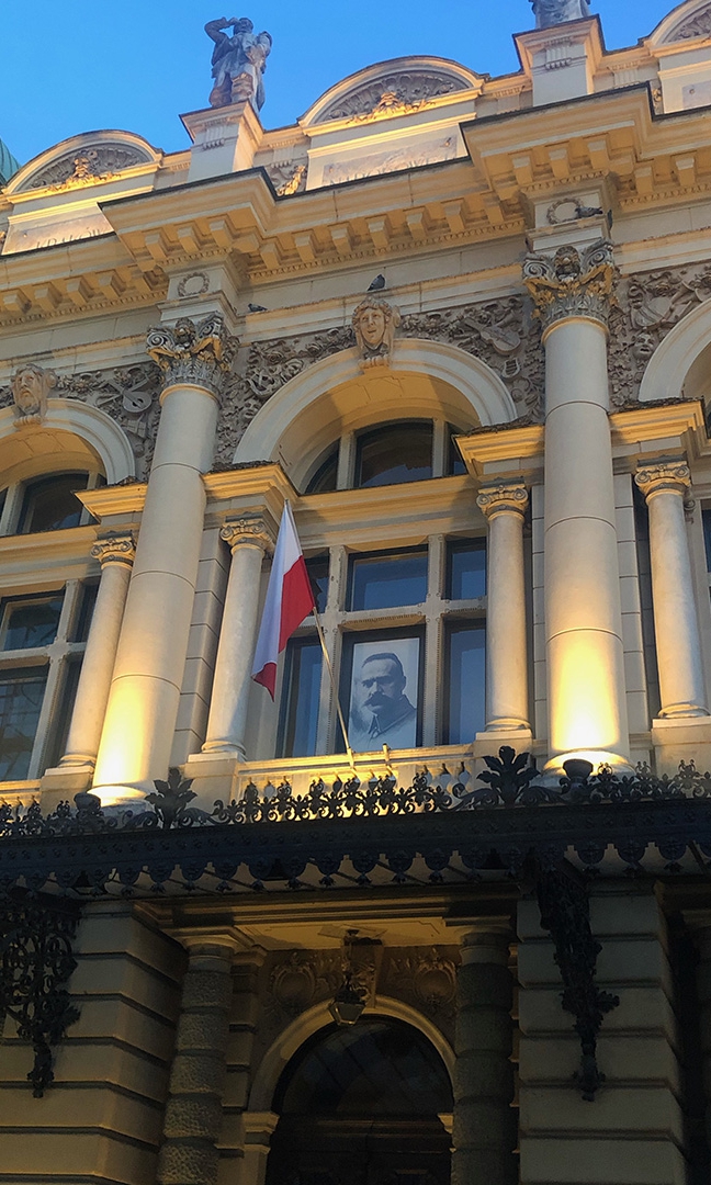 Zdjęcie – górna część frontonu budynku Teatru Słowackiego. W oknie budynku zawieszony portret Marszałka Piłsudskiego. Obok nabita na maszt flaga Polski w barwach biało-czerwonych.
