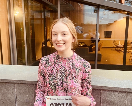Na zdjęciu młoda dziewczyna w różowej, kwiecistej koszuli i spodniach. W dłoniach trzyma kartkę z numerem biegacza. Jest uśmiechnięta. Za nią przestrzeń konferencyjna budynku Małopolskiego Ogrodu Sztuki.