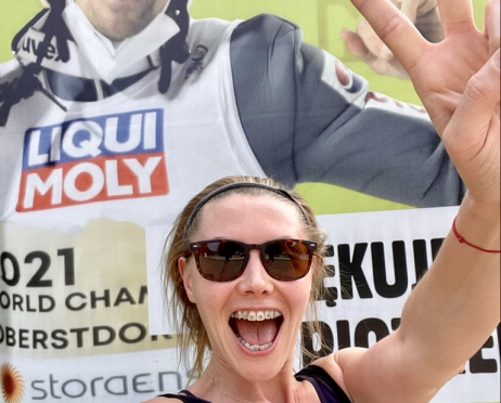 Na zdjęciu młoda kobieta pozuje na tle baneru reklamowego. Jest roześmiana, ma aparat na zębach. Nosi okulary przeciwsłoneczne. Jedna z rąk unosi w górę. Baner reklamowy przedstawia jednego z polskich skoczków narciarskich.