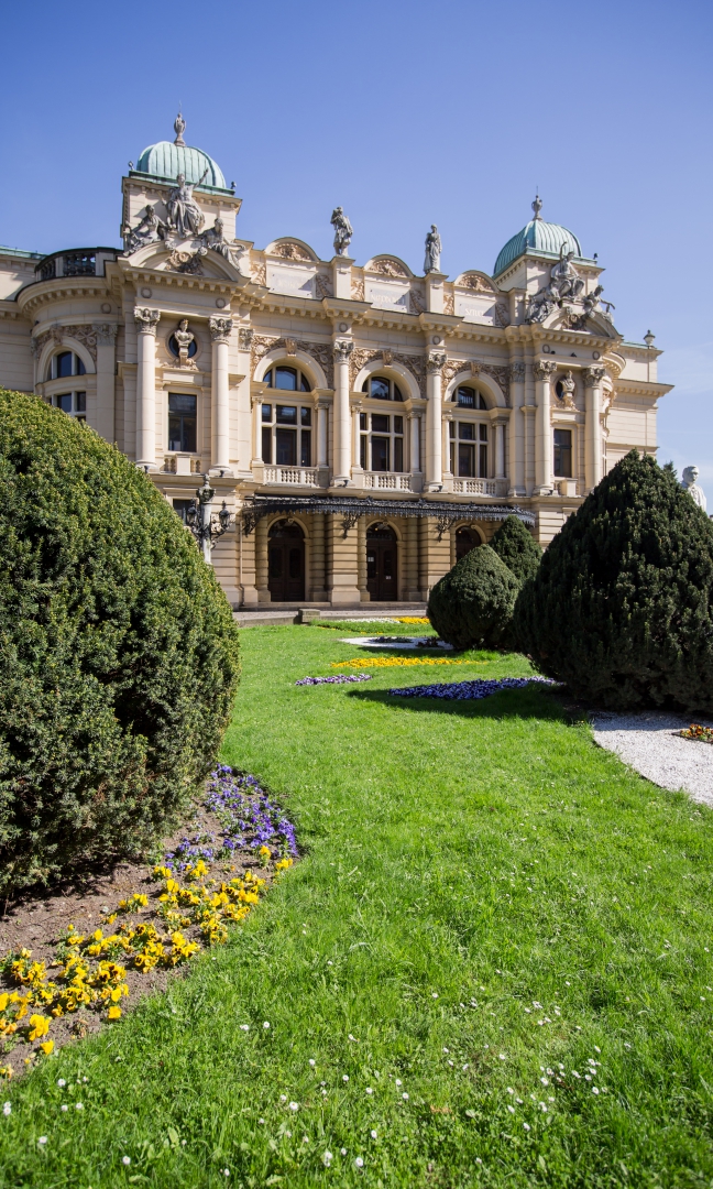 Zdjęcie przedstawia budynek Teatru im. Juliusza Słowackiego w Krakowie. Fasadę zdobią rzeźby. Do budynku prowadzą trzy wejścia. Nad nimi trzy duże okna.  Przed budynkiem zielone krzewy.