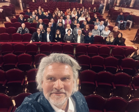 Zdjęcie robione ze sceny przez Dyrektora Teatru. Jest to selfie. W oddali widownia. Na bordowych fotelach siedzi grupa osób. Kobiety i mężczyźni. Uśmiechają się. Patrzą prosto przed siebie.