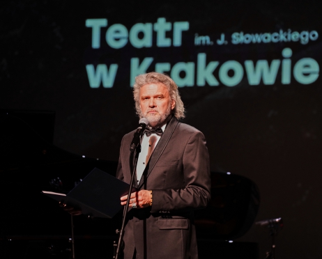 Na zdjęciu dyrektor Teatru Słowackiego Krzysztof Głuchowski. Ubrany w czarny smoking i białą koszulę, stoi przed mikrofonem. W tle ekran z wyświetlony logotypem Teatru w Krakowie.
