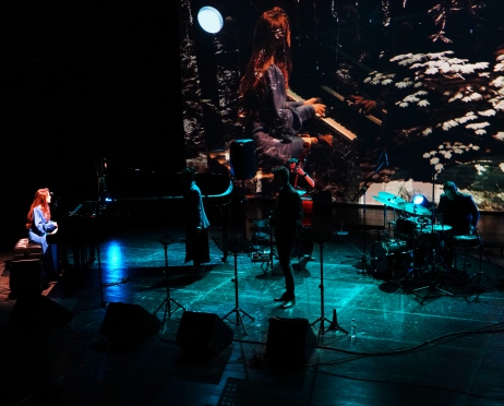 Zdjęcie ciemnej sceny z bardzo słabo oświetlonymi punktami. Na scenie mikrofony, perkusja, fortepian  i cztery osoby. W tle projekcja na ekranie - zbliżenie kobiety która gra na fortepianie.