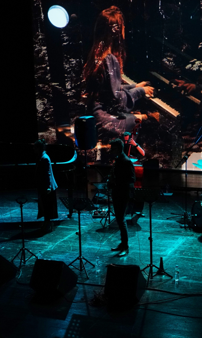Zdjęcie ciemnej sceny z bardzo słabo oświetlonymi punktami. Na scenie mikrofony, perkusja, fortepian  i cztery osoby. W tle projekcja na ekranie - zbliżenie kobiety która gra na fortepianie. .