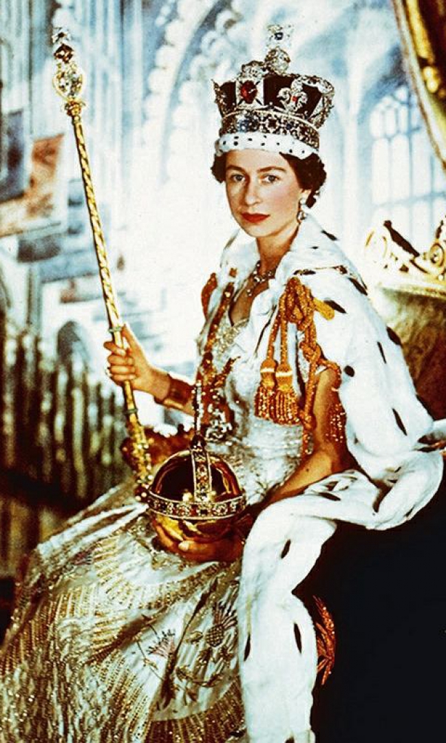 Zdjęcie przedstawia królową siedzącą na tronie. Ubrana jest w złotą suknię, na ramionach ma purpurowy płaszcz a na głowie ma koronę. W dłoniach trzyma berło i jabłko. W tle kolumny zdobiące wnętrze budynku.