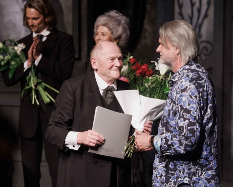 Na zdjęciu dyrektor Teatru Krzysztof Głuchowski ubrany w błękitną koszulę w kwiaty obok starszego mężczyzny ubranego w smoking. Uśmiechają się. Mężczyzna trzyma w dłoniach bukiet kwiatów i tekturową teczkę. Z tyłu stoi starsza kobieta i młody mężczyzna. Obydwoje trzymają bukiety kwiatów, klaszczą.
