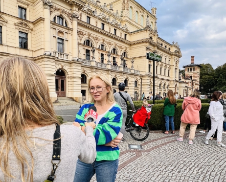 Na pierwszym planie kobieta w zielonym swetrze udziela wywiadu. Za nią budynek Teatru im. Juliusza Słowackiego w Krakowie. Budynek jest koloru żółtego. Poszczególne piętra elewacji ozdobione są niewielkimi rzeźbami. Na placu przed budynkiem gromadzą się ludzie. Niebo jest pochmurne.