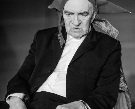 Czarno-białe zdjęcie Jerzego Treli. Aktor jest starszy, siedzi na krześle niechlujnie. Ma rozpiętą marynarkę. Na głowie ma czapkę błazna. Jest poważny, patrzy w podłogę.