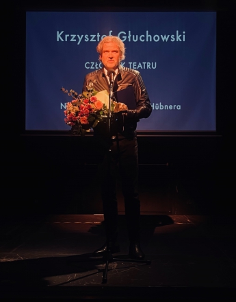 Na zdjęciu Krzysztof Głuchowski Dyrektor Teatru Słowackiego. Stoi na scenie przed mikrofonem, w dłoniach trzyma kwiaty i dyplom. Ubrany w skórzaną, czarną kurtkę. Za nim duży ekran na którym wyświetlone jest jego nazwisko i nazwa nagrody którą zasłania. Scena jest ciemna, światło pada tylko na niego.