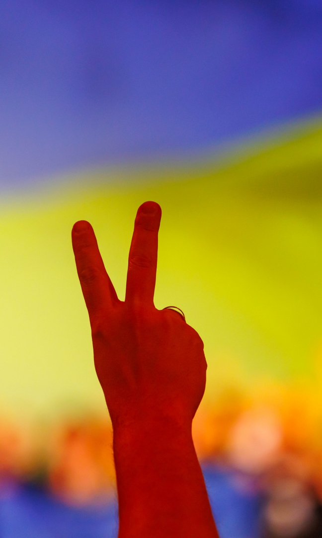 Na tle niebiesko-żółtego tła, sugerującego flagę Ukrainy jest uniesiona w górę dłoń z wysuniętymi dwoma palcami na znak pokoju. Dłoń jest w czerwonej poświacie.