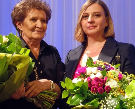Na zdjęciu dwie kobiety- starsza i w średnim wieku. Obydwie ubrane w ciemne marynarki, w dłoniach trzymają bukiety kwiatów. Jedna z kobiet dumnie patrzy w obiektyw. W tle błękitna kotara.