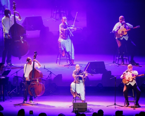 Na scenie trzech muzyków: Mężczyzna grający na kontrabasie, w środku na wysokim krześle siedzi kobieta, gra na skrzypcach, śpiewa do mikrofonu. po prawej stronie mężczyzna, siedzi na wysokim krześle, ma zawieszoną na szyi gitarę, rękoma poprawia mikrofon na stojaku. po lewej stronie zdjęcia w tle perkusista. Za nimi na wielkim ekranie powiększone i powielone postaci muzyków. Na scenie niebiesko-fioletowe światło.