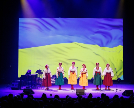 Na scenie w rzędzie stoi sześć kobiet w kolorowych strojach ludowych. Mają białe koszule i spódnice do kostek w różnych kolorach oraz czerwone trzewiki. Na głowie obwiązane czerwone chusty, na szyi czerwone korale. W tle za nimi wielki ekran na którym wyświetlona jest flaga Ukrainy. Po lewej stronie stoi fortepian i perkusja. U dołu zdjęcia cienie głów osób siedzących na widowni.