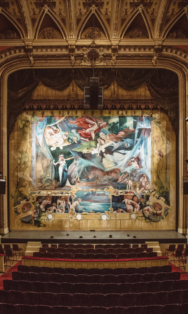 Ujęcie na scenę Teatru Słowackiego z opuszczoną kurtyną Stanisława Wyspiańskiego. Kurtyna jest malowana, kolorowa, przedstawia abstrakcyjny obraz Krakowa widziany oczyma Wyspiańskiego.