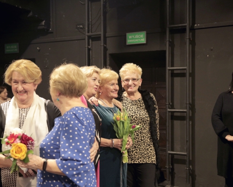 Zdjęcie przedstawia pięć kobiet w starszym wieku. Stoją w grupie. Rozmawiają ze sobą. Trzy z nich pozują do zdjęcia. Szeroko się uśmiechają. Jedna z kobiet trzyma bukiet kwiatów. Po prawej stronie dwie kobiety które patrzą w kierunku pozujących kobiet do zdjęcia. Wszystkie osoby na zdjęciu ubrane są elegancko. Tło jest czarne.