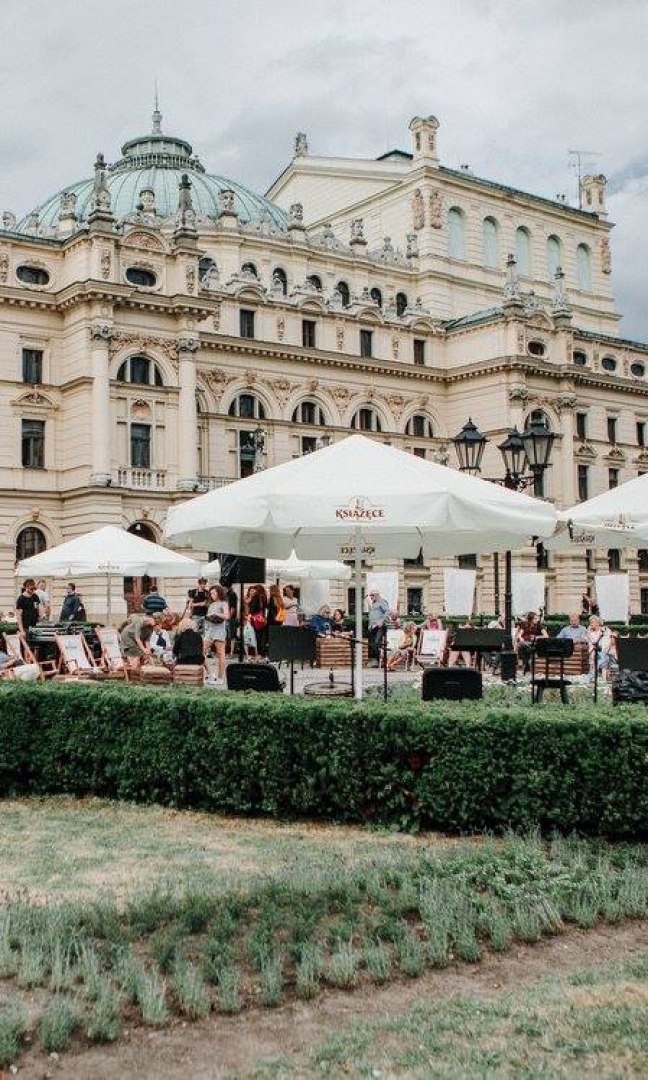 Zdjęcie budynku Teatru Słowackiego od strony pl. Św Ducha. Przed budynkiem rozłożone duże parasole kawiarniane w kolorze kremowym z napisem Książęce oraz leżaki. Pomiędzy nimi chodzą ludzie w letnich strojach.