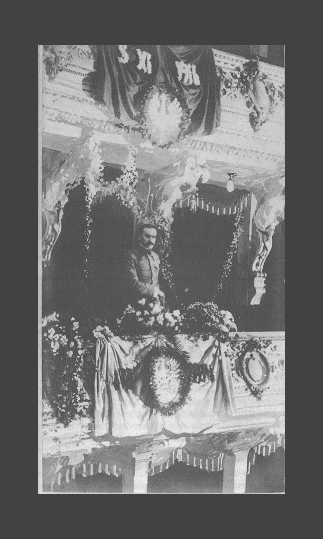 Czarno-biała fotografia przedstawia Józefa Piłsudskiego stojącego w loży teatralnej. Mężczyzna ubrany jest w mundur. Loża teatralna jest bogato przystrojona bukietami kwiatów i girlandami kwiatowymi. Na lożach dodatkowo zamontowano polskie godło.