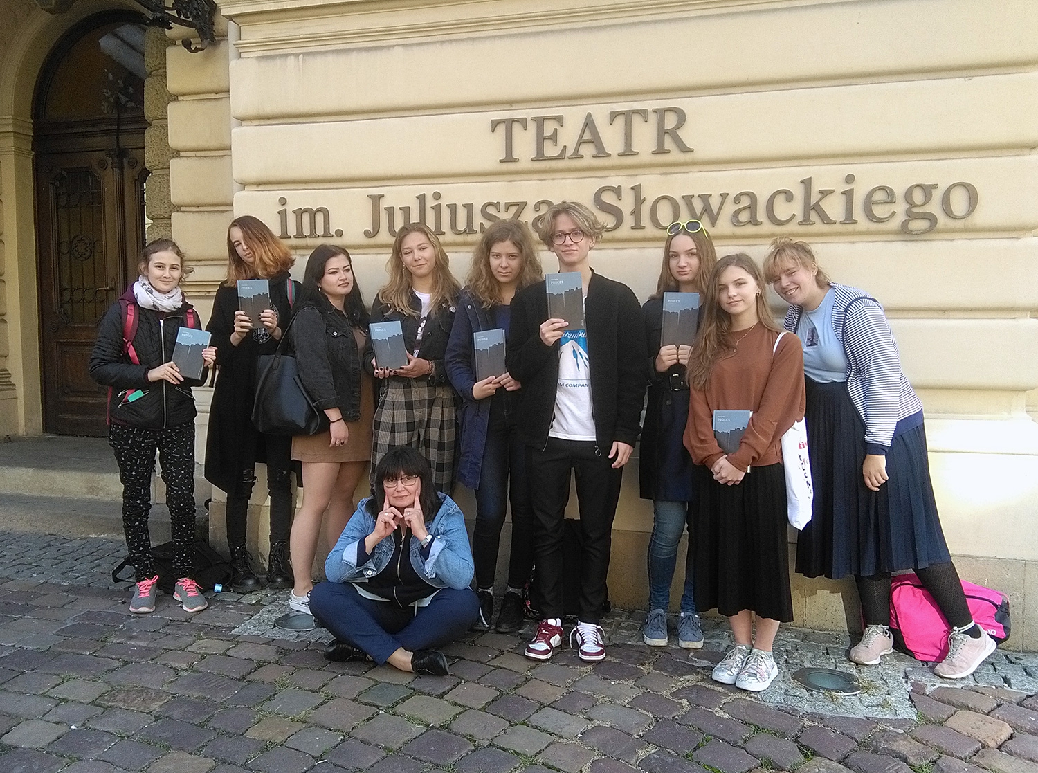 Podjazd przed wejściem głównym do Teatru im. J. Słowackiego. Grupa młodzieży stoi. Większość trzyma egzemplarze "Procesu" Franza Kafki. Przed grupą młodzieży kuca nauczycielka.