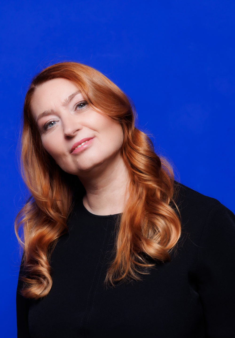 Portret Bożeny Sowy, kobiety w średnim wieku, o długich, falowanych rudych włosach, jasnej cerze i jasnoniebieskich oczach. Kobieta lekko przechyla głowę, patrzy w obiektyw i delikatnie się uśmiecha. Ma na sobie czarną bluzkę z przeszyciem na środku. Kobieta stoi na intensywnie niebieskim tle.