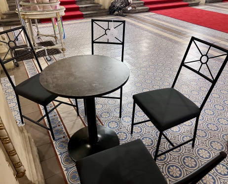 Na zdjęciu zestaw stół i cztery krzesła. Stół jest czarny i okrągły. Wokół stoją cztery klasyczne metalowe krzesła z szarymi poduszkami w kształcie prostokąta. Zestaw stoi na posadzce z mozaikowym wzorem w stylu portugalskim. W tle schody przykryte czerwonym dywanem.