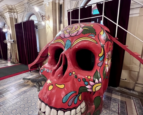 Wnętrze westybulu Teatru Słowackiego. Mozaikowa posadzka, bogate zdobienia na ścianach, kotary w drzwiach. Na środku pomieszczenia stoi ogromna czerwona czaszka wymalowana w meksykańskie wzory. Ma wielkie białe zęby. Z jednej strony wejście na czaszkę jest po schodach z balustradą, z drugiej strony czerwona zjeżdżalnia. Obok czaszki leży mały, sztuczny krokodyl.