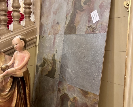 O beżową ścianę na korytarzu teatru stoi oparta wielka kamienna płyta, złożona z kwadratów, gdzie każdy z nich ma inny wzór i kolor. Na płycie naklejona jest kartka. Obok widoczny fragment figury - dzieciątka, które jest nagie, prawdopodobnie jest to figura dzieciątka Jezus na rękach Maryi. W tle tralki na schodach w beżowym kolorze.