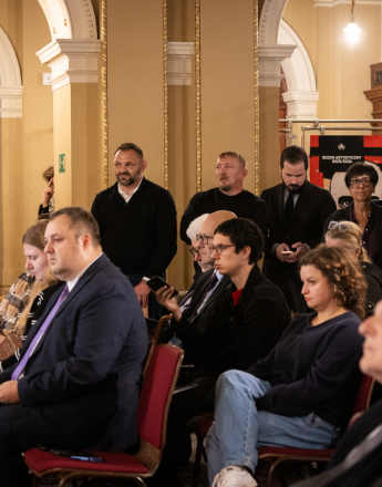 W reprezentacyjnym westybulu Teatru imienia Juliusza Słowackiego w Krakowie zgromadzeni są uczestnicy konferencji prasowej oraz pracownicy teatru, w tym osoby z dyrekcji, kadry kierowniczej i zespołu teatru. W centrum i po lewej stronie obrazu w kilku rzędach siedzą uczestnicy konferencji. W pierwszym planie po lewej stronie siedzi mężczyzna w granatowym garniturze, białej koszuli i fioletowym krawacie. Za nim widoczny jest mężczyzna w okularach, ubrany w czarną marynarkę i białą koszulę, trzymający telefon. Obok niego siedzi kobieta w czarnym stroju z czerwonym akcentem przy szyi. W centrum siedzi kobieta z kręconymi, brązowymi włosami, ubrana w ciemny sweter i jasne jeansy. W dalszej części sali, za siedzącymi uczestnikami, stoi grupa pracowników i osób związanych z teatrem. W tle znajdują się charakterystyczne dla zabytkowego wnętrza teatru wysokie, jasne arkady, dekoracyjne kolumny, złocone detale architektoniczne oraz drewniane elementy. Po lewej stronie stoi plansza przedstawiająca budynki i architekturę Krakowa. W centrum, za grupą osób, znajduje się plakat promujący sezon artystyczny 2025/2026. Po prawej stronie widoczna jest profesjonalna kamera skierowana w stronę uczestników konferencji. Cała grupa jest zwrócona w stronę wydarzenia odbywającego się poza obszarem widocznym na obrazie.