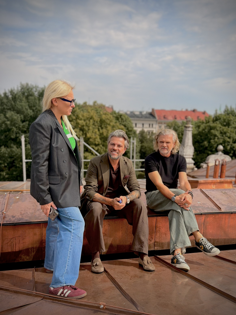 Trzy osoby stoją na dachu Teatru imienia Juliusza Słowackiego w Krakowie. Po lewej stoi zastępczyni dyrektora do spraw wizerunku i komunikacji Edyta Sotowska-Śmiłek, ubrana w ciemną marynarkę, zielony top i jasnoniebieskie jeansy. Pośrodku siedzi Oliwier Janiak, ubrany w brązową marynarkę, ciemną koszulę i spodnie. Po prawej siedzi dyrektor teatru Krzysztof Głuchowski, w czarnej koszulce i jasnych spodniach. Edyta Sotowska-Śmiłek spogląda na siedzących mężczyzn, a mężczyźni patrzą przed siebie. Wszyscy znajdują się na miedzianym dachu. W tle widać fragmenty dachu teatru, zieleń drzew oraz zabudowę Krakowa. 