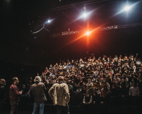 Sala widowiskowa, zdjęcie zrobione ze sceny w kierunku widowni. W rzędach krzeseł ustawionych kaskadowo siedzi publiczność. Patrzą na scenę, na której w rzędzie stoją czterej mężczyźni. Mężczyźni są sfotografowani od tyłu. Z góry na publiczność skierowany jest reflektor,który oświetla widownię ciepłym, pomarańczowym światłem.