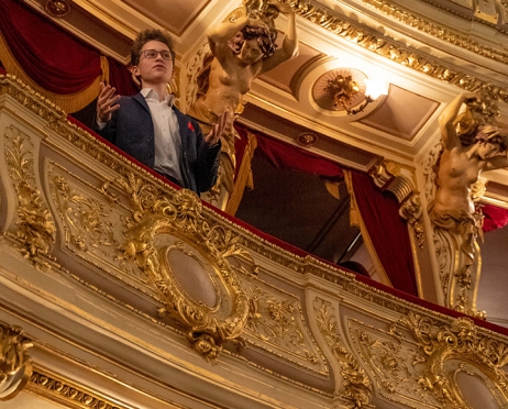 Na zdjęciu w centralnej części praktycznie na całej powierzchni loża cesarska. Bogato zdobiona. W loży stoi młody chłopak około siedemnastu lat. Patrzy w kierunku widowni. Ubrany jest w garnitur i białą koszulę. Ręce ma podniesione lekko w górę. Tłem jest fragment złoto zdobionej loży.