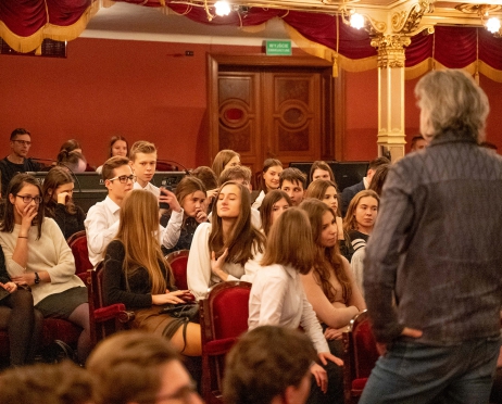 Na zdjęciu fragment widowni. W prawym dolnym rogu dyrektor Teatru. Patrzy w stronę młodych ludzi,. Podpiera się za boki. Młodzież patrzy w jego stronę. niektórzy mają rozproszone spojrzenie. NA widowni panuje chaos.  Tło jest czerwone.