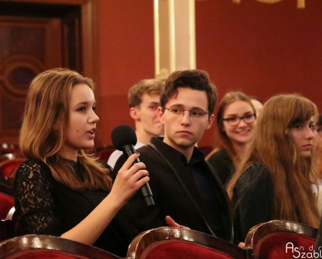 Na zdjęciu fragment widowni. W centralnej części siedzi młoda dziewczyna na czerwonym fotelu. Siedzi bokiem. W ręku trzyma mikrofon. Coś mówi. Obok chłopak, patrzy w jej stronę. Obok niego młoda dziewczyna patrzy w stronę sceny. W tle rząd widowni. Twarze osób skierowane na dziewczynę która mówi.