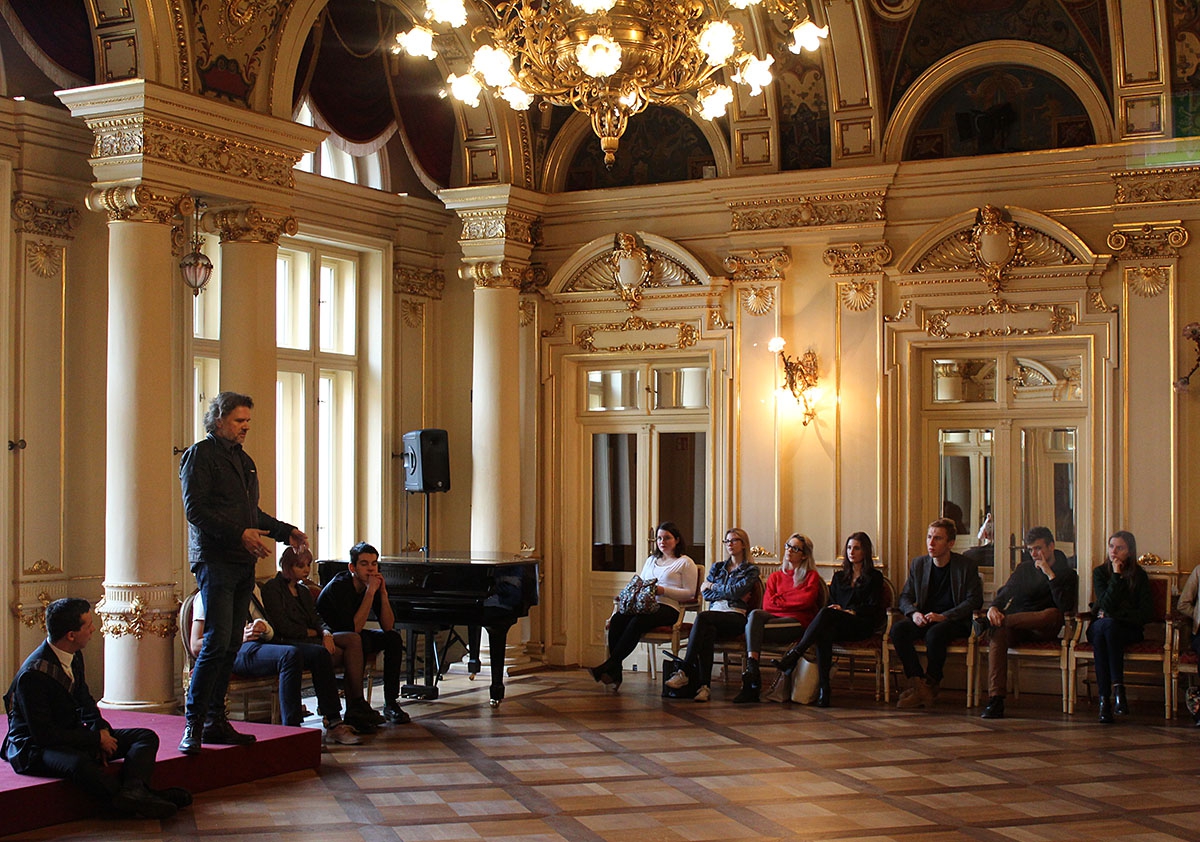 Pięknie zdobione wnętrze foyer teatru. Grupa ludzi siedzi na ozdobnych krzesłach ustawionych przy ścianach. Na podwyższeniu (mała scena) dyrektor teatru mówi do młodzieży.