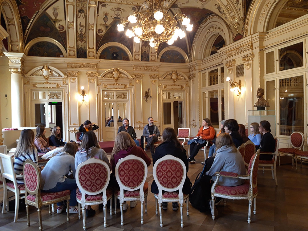 Bogato zdobione wnętrze foyer, z pięknym, dużym  żyrandolem. Na pięknych krzesłach, w kręgu siedzi młodzież z dyrektorem teatru i dramaturgiem. Z prawej strony - popiersie Stanisława Wyspiańskiego.