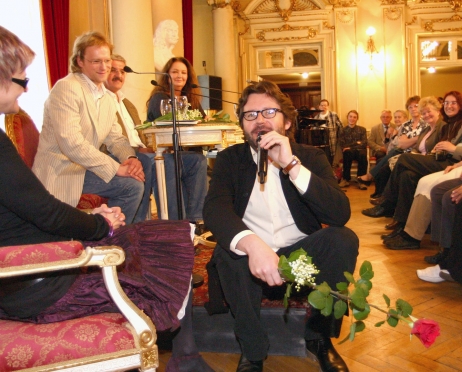 Foyer Teatru. Na małym podwyższeniu siedzą na krzesłach: Maciej Stuhr, Marek Niedźwiecki i Anna Dymna - są widoczni od boku. Spoglądają w kierunku siedzącego do nich tyłem na brzegu małej sceny Grzegorza Turnaua. W lewej ręce trzyma mikrofon, w drugiej kwiaty - czerwoną różę i mały bukiet konwalii. Jest zwrócony w kierunku kobiety siedzącej na ozdobnym fotelu. Po prawej stronie siedzą widzowie. Wszyscy są ubrani elegancko.