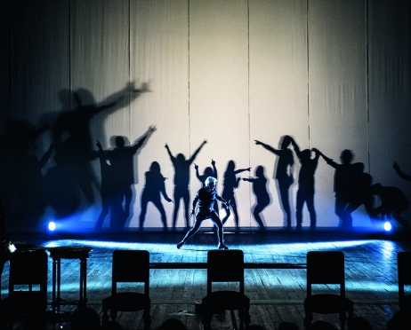 Zdjęcie przedstawia scenę. Na scenie na pierwszym planie rząd pustych krzeseł. Za nim tańcząca kobieta ubrana w czarny kombinezon. Kobietę oświetlają dwa niebieskie reflektory od dołu. Tłem jest białe płótno na którym są cienie tańczących osób w rzędzie.