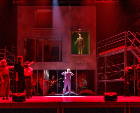 Scena teatru oblana czerwonym światłem. Na niej konstrukcja przypominająca piętrowy budynek. Na ścianie neonowy napis “mięso” po prawej i lewej stronie budynku stoją rusztowania. Na parterze za szybą siedzą muzycy przy instrumentach W jednym z okien budynku na piętrze jest zapalone zielone światło, stoi w nim mężczyzna w zielony , wojskowym garniturze. Przed budynkiem na środku sceny stoi młody mężczyzna w fioletowym garniturze, uśmiecha się, dłonie trzyma pod brodą. Wokół niego kobiety stojące tyłem do obiektywu, wyciągają dłonie w jego stronę.