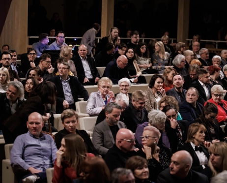 Zdjęcie na oświetloną salę widowiskową. Krzesła na widowni są białe i kwadratowe. Sala jest pełna widzów, ludzie rozmawiają, uśmiechają się. Na widowni m.in Danuta Wałęsa, Krzysztof Głuchowski i Henryka Krzywonos.