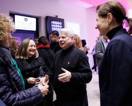 Na zdjęciu luźne spotkanie i rozmowy osób. Na pierwszym planie Bogdan Borusewicz witający się z kobietą, Towarzyszące im pozostałe dwie kobiety szeroko uśmiechają się. W tle więcej osób, ściana podświetlona różowym kolorem.