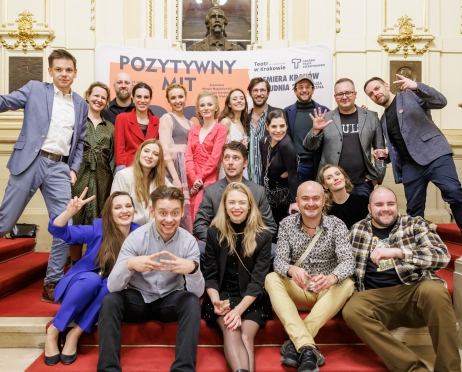 Grupa osób jest ustawiona do zdjęcia w rzędach jak do szkolnej fotografii. Uśmiechają się. Za nimi ścianka premierowa z napisem Pozytywny mit. W tle zdobione ściany i czarne rzeźbione popiersie.