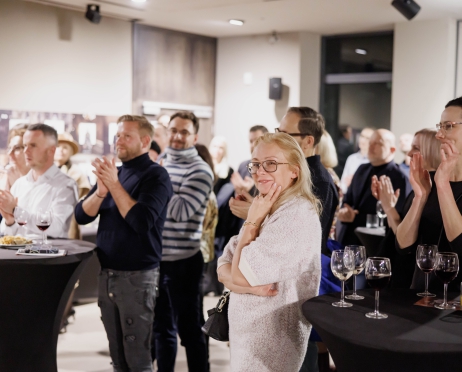 Na zdjęciu grupa ludzi. Stoją mniejszymi grupami przy wysokich okrągłych stolikach. Na stolikach kieliszki z winem. Wszyscy patrzą w jednym kierunku są skupieni. Niektórzy klaszczą. W centralnej części na środku kobieta. Jest uśmiechnięta. Rękę opiera o brodę. Ubrana jest w jasny sweter. Tło jest białe