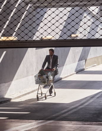 Na zdjęciu mężczyzna w średnim wieku prowadzi wózek z zakupami do podziemnego garażu. Wchodzi do garażu od strony ulicy, drogą dla samochodów. Nad wjazdem znajduje się do połowy opuszczona brama. Mężczyzna ma na sobie popielaty garnitur i białą koszulę.