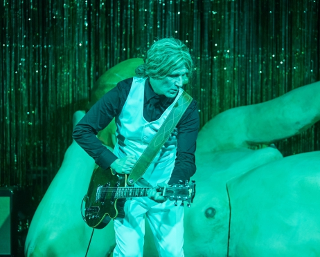 Na fotografii mężczyzna w białych spodniach i kamizelce gra na gitarze. Ma długie, jasne włosy spięte w kucyk. Spogląda w dół. Za nim ustawiona jest duża rzeźba przedstawiająca nagiego mężczyznę. W tle wiszą lśniące wstążki. Na scenę pada zielone światło.