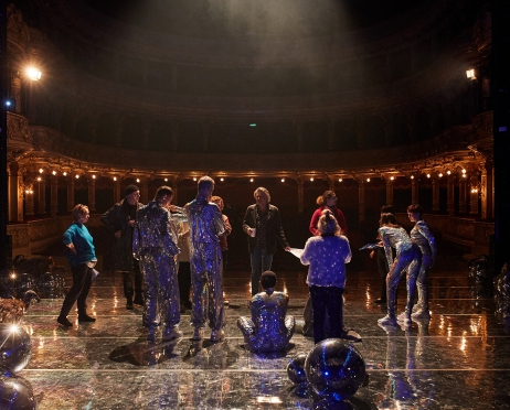 Zdjęcie wykonane na Dużej Scenie Teatru. Na tle widowni stoi kilkunastoosobowa grupa. Wśród nich znajdują się aktorzy w srebrnych kostiumach oraz twórcy spektaklu i pracownicy teatru ubrani współcześnie. Po bokach sceny ułożone na sobie srebrne kule. W tle widownia i loże.