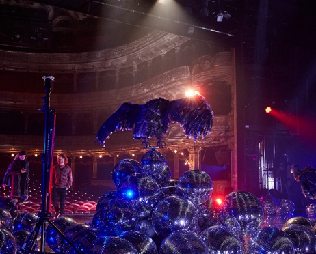 Zdjęcie wykonane na Dużej Scenie Teatru.  W centralnym miejscu na stercie dyskotekowych kul ustawiono wypchanego sępa z rozpostartymi skrzydłami. W tle widownia i loże. W tle dwoje mężczyzn ubranych współcześnie.