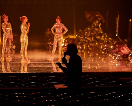 Zdjęcie Dużej Sceny i widowni Teatru. Na scenie stoi w nieładzie kilka osób w błyszczących kombinezonach. W tle ułożone na sobie kule dyskotekowe. Między rzędami w półmroku sylwetka mężczyzny z mikrofonem w dłoni.