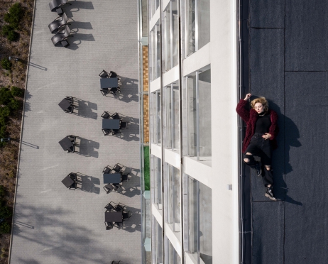 Na zdjęciu kobieta ubrana w czarne spodnie, golf i bordowe, sztuczne futerko. Leży na dachu budynku. Ma zamknięte oczy. Przed budynkiem stoliki i krzesła. Zdjęcie zrobione z góry.