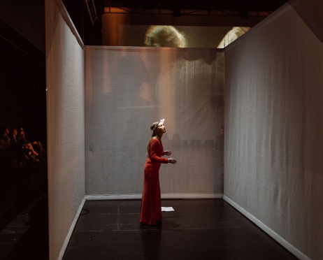 W małym pomieszczeniu złożonym z trzech niebieskich ścian stoi zdezorientowana kobieta. Jest ubrana w czerwoną sukienkę, twarz ma oświetloną małymi lampkami, które są wmontowane w opaskę na jej głowie.