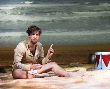 Na plaży na piasku siedzi młody mężczyzna w skarpetach, koszuli i krótkich spodenkach. Za nim jest morze. Mężczyzna trzyma uniesiony w górę palec wskazujący. Obok niego w piasku leży bęben.
